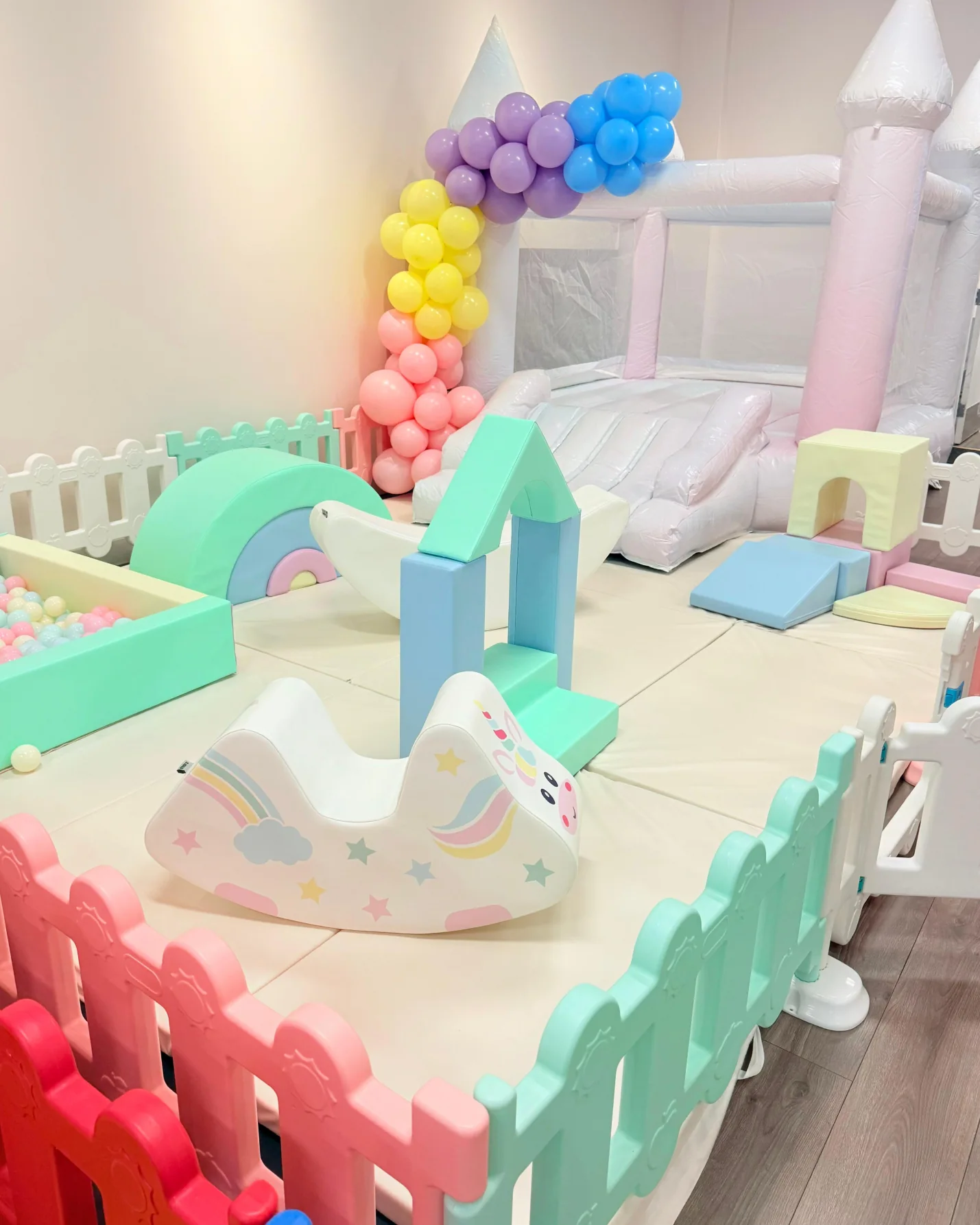 Pastel soft play rental with white bounce castle slide unicorn rocker ball pit building blocks and rainbow balloon garland for indoor toddler party in El Paso